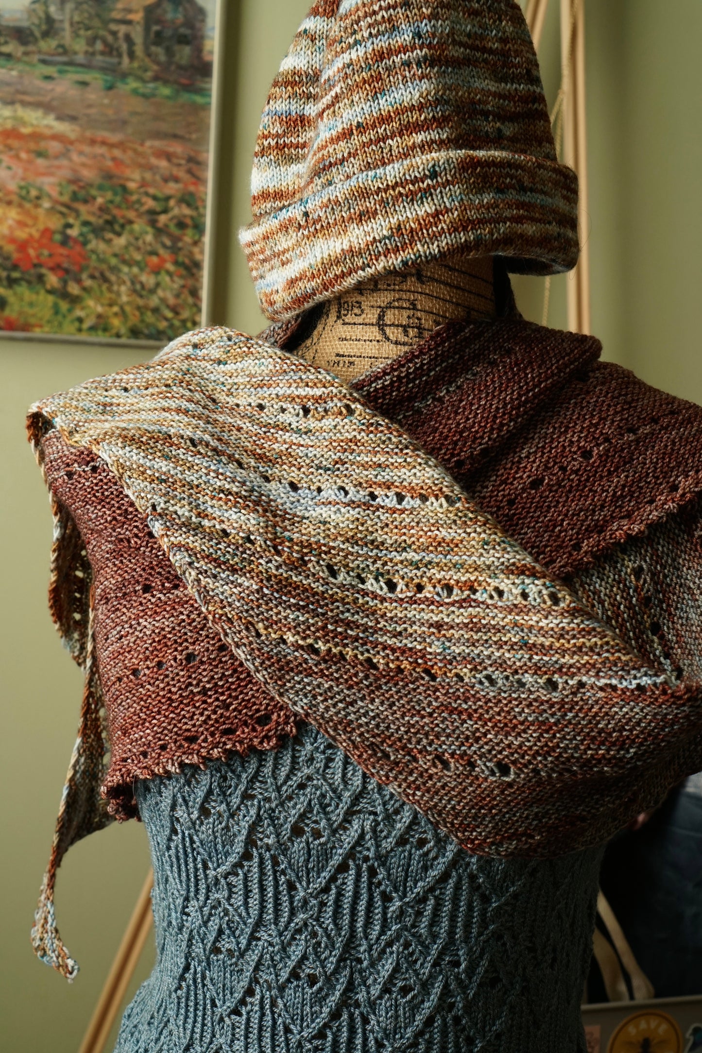 Mannequin wearing a faded garter knit shawl and hat in earth tones with a green blurred background.