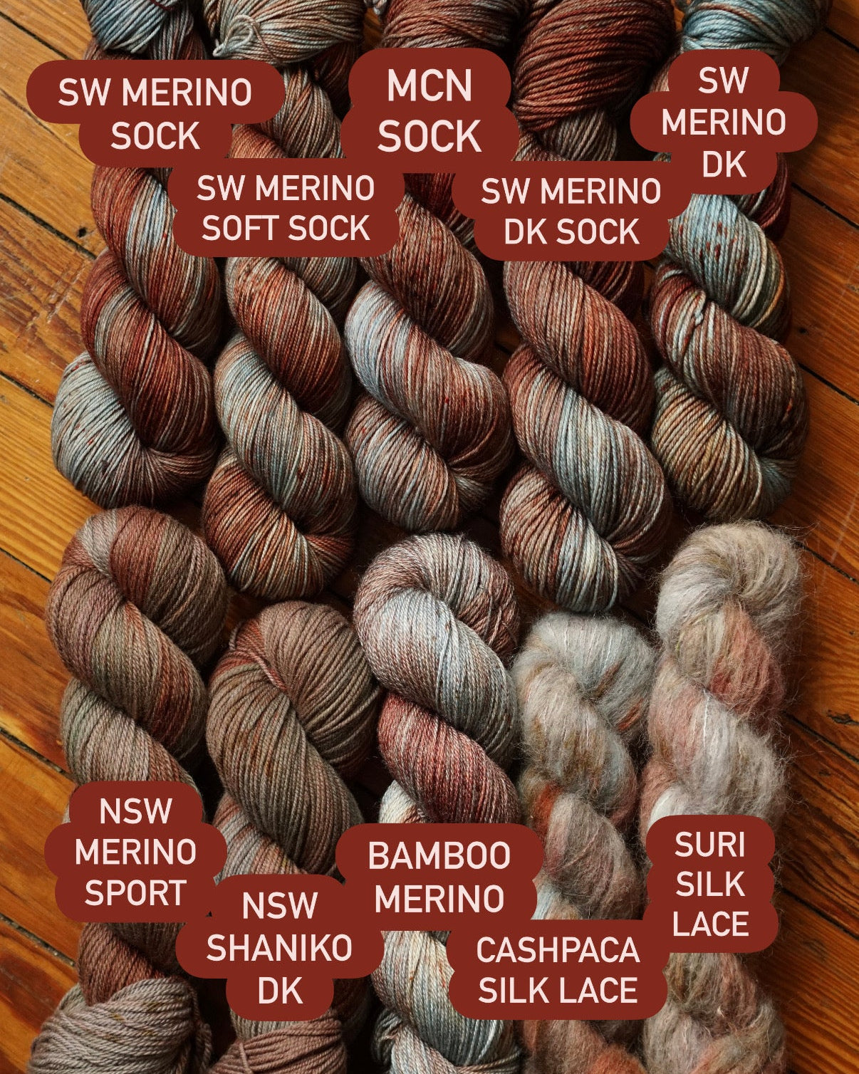 Multicolored red, brown and teal yarn skeins in a variety of fiber blends arranged neatly on a wooden floor, labeled by fiber blend. 