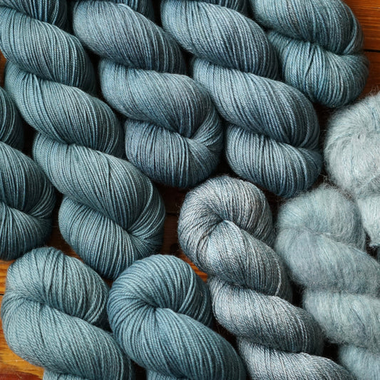 Skeins of slate teal grey skeins in a variety of fiber blends arranged at a slant on a wooden surface.