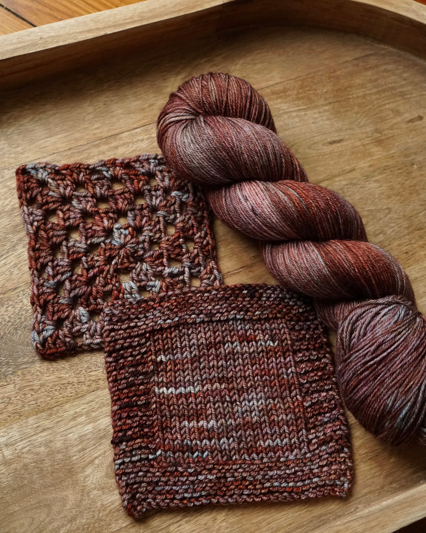 Knitted swatches and a skein of red brown yarn with slate teal pops of color on a wooden tray
