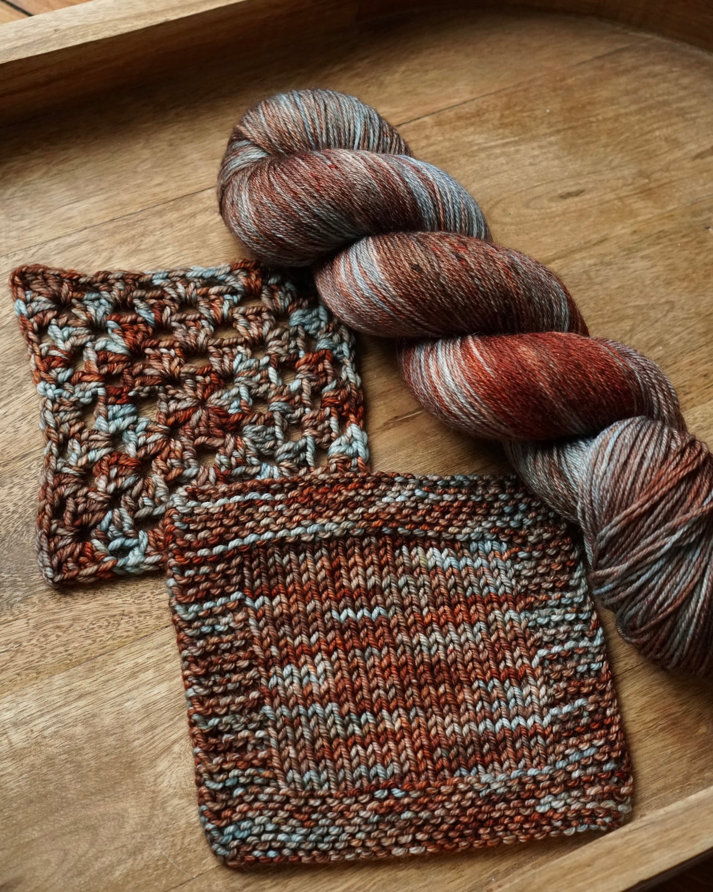 Knitted swatches and a skein of red,brown and teal yarn on a wooden tray