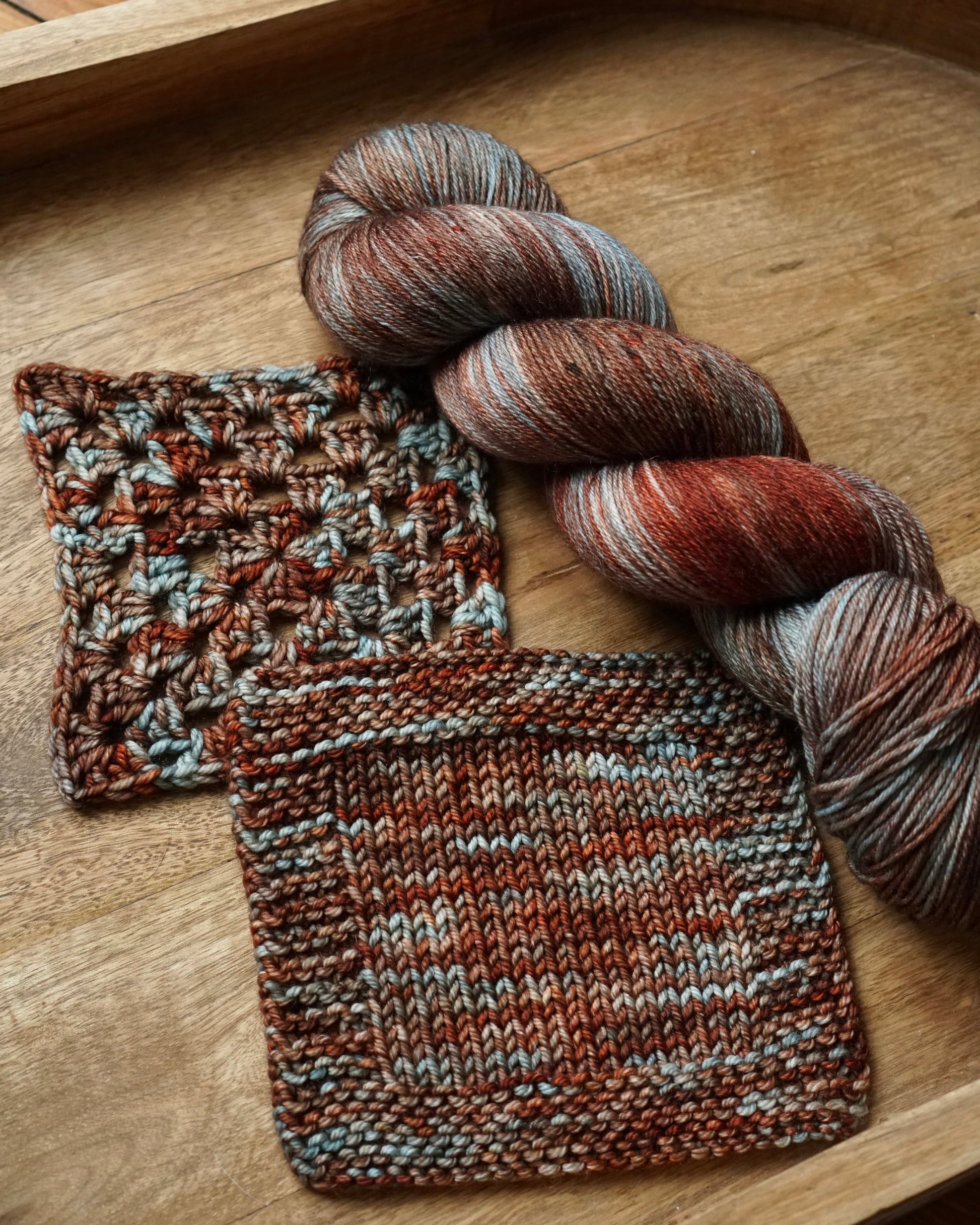 Knitted swatches and a skein of red,brown and teal yarn on a wooden tray