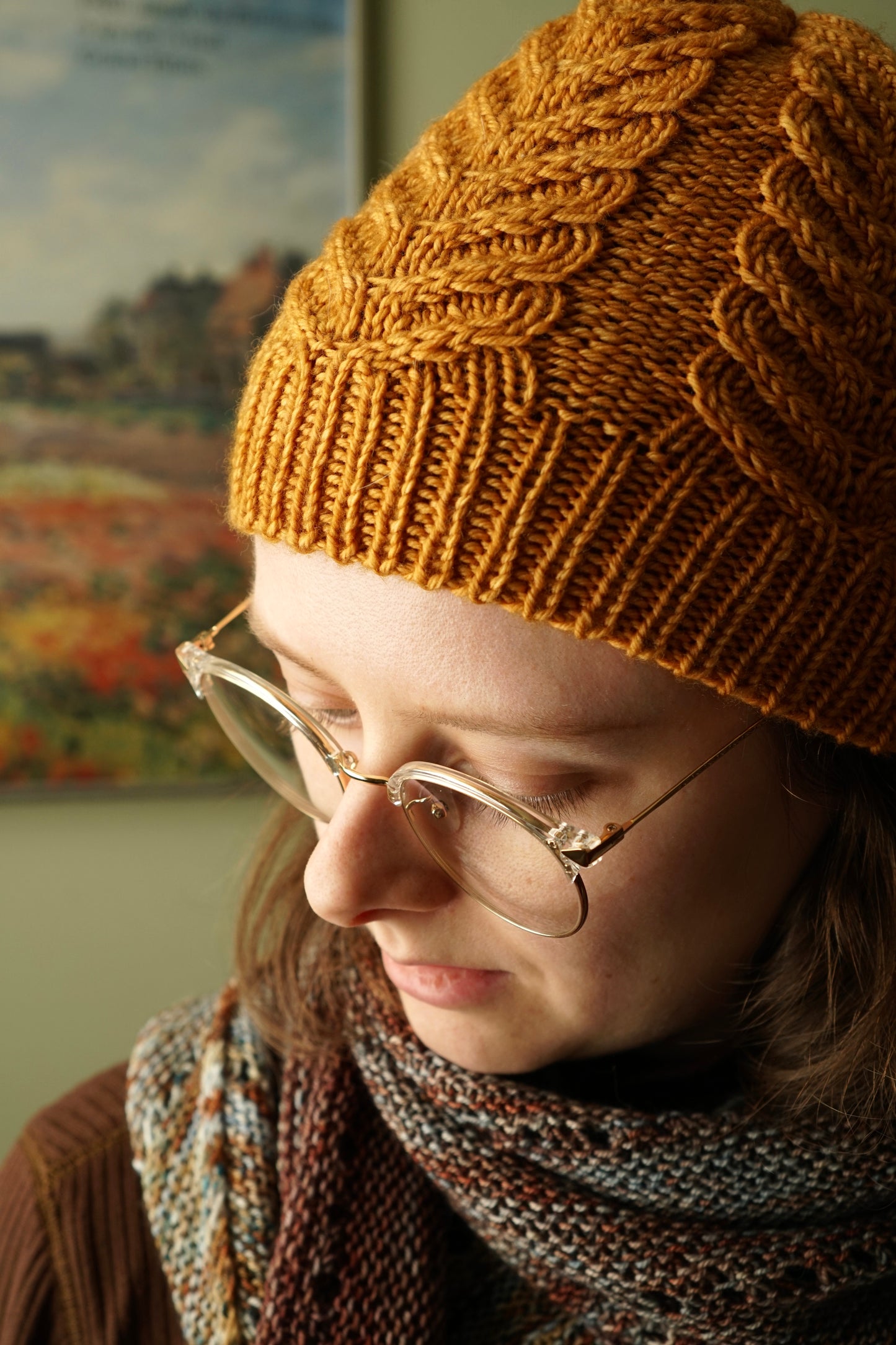 Person wearing a cable-knit golden yellow hat and glasses, with a blurred painting in the background.