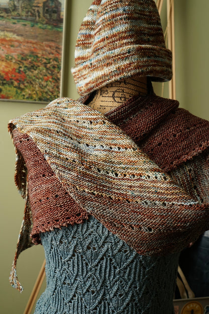 Mannequin wearing a faded garter knit shawl and hat in earth tones with a green blurred background.
